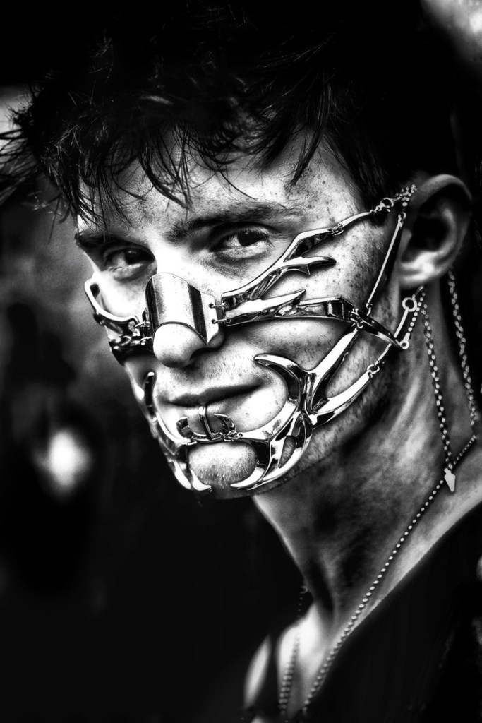 A close-up portrait of a person with an elaborate metallic facial mask, featuring intricate designs and a striking expression, set against a blurred dark background.